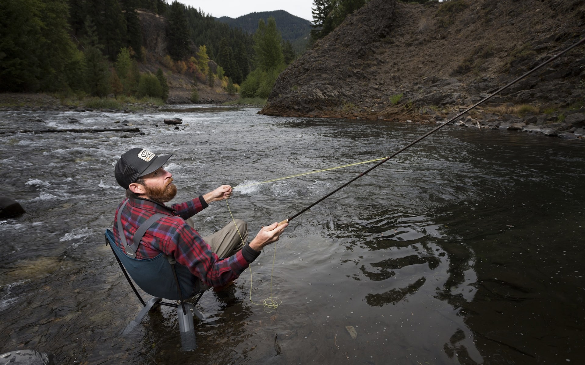 Man in outdoor outfit sitting in dark blue Therm-a-Rest Quadra camp chair by ITO Design in a river, holding a fishing pole