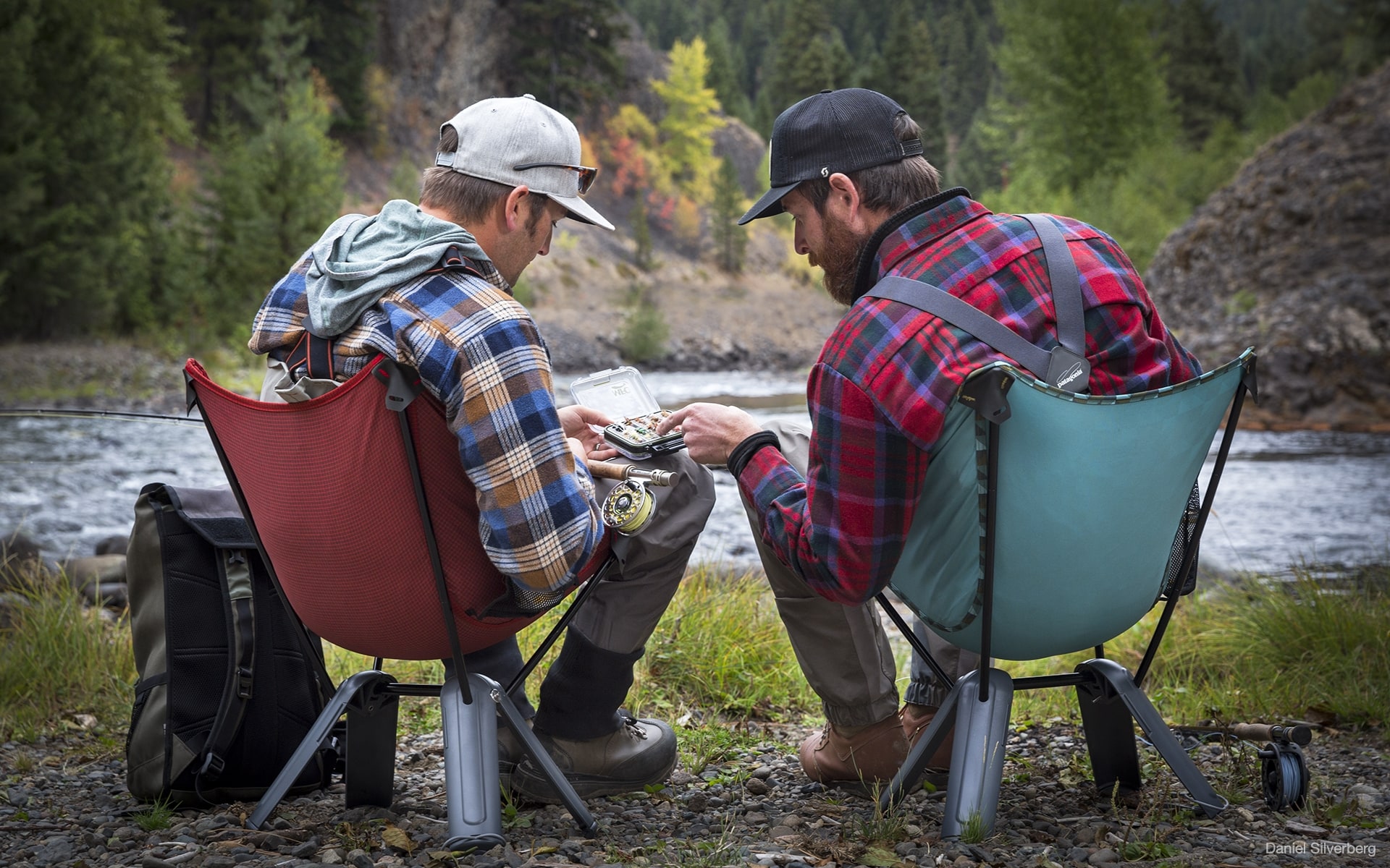 Zwei Männer in Outdoor-Kleidung sitzen auf roten und grünen Therm-a-Rest Quadra Camping-Stühlen von ITO Design an einem Fluss und sehen sich Angelköder an