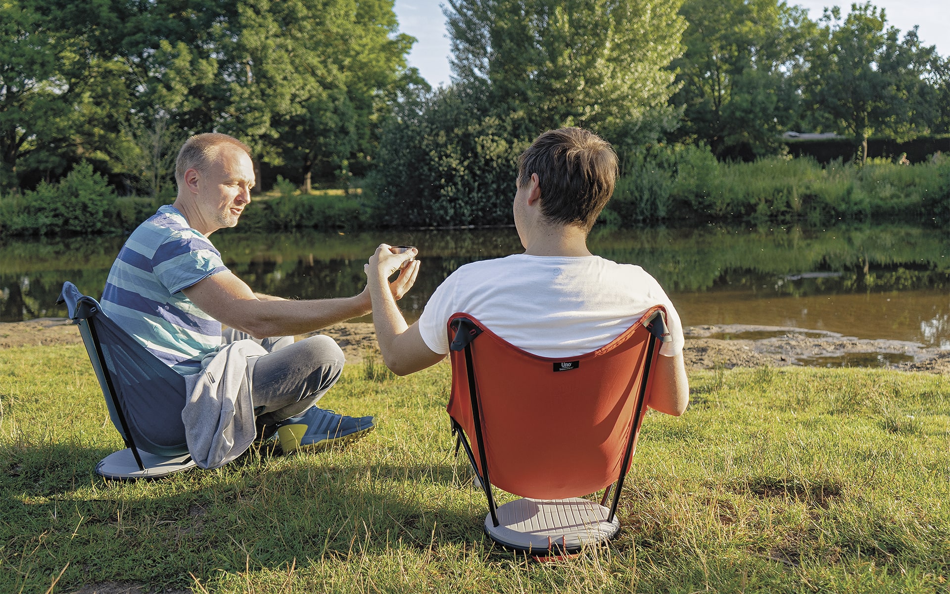 Zwei Männer in Freizeitkleidung sitzen in einem blauen und einem roten Therm-a-Rest Uno Outdoor-Stuhl von ITO Design neben einem Fluss, der eine reicht dem anderen einen Becher