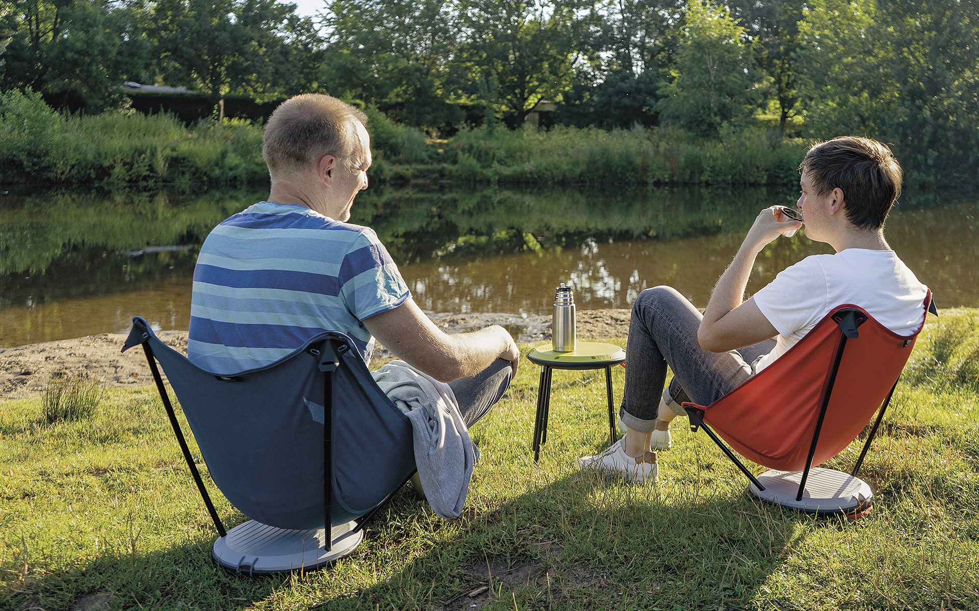 Two men sitting in blue and red Therm-a-Rest Uno outdoor chairs by ITO Design near river, using a collapsed Therm-a-Rest Uno outdoor chair as a side table