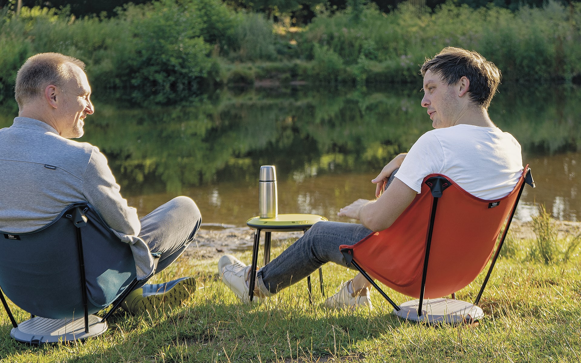 Zwei Männer sitzen in Therm-a-Rest Uno Outdoor-Stühlen von ITO Design neben einem Fluss, nutzen einen zusammengeklappten Therm-a-Rest Uno Outdoor-Stuhl als Beistelltisch und unterhalten sich